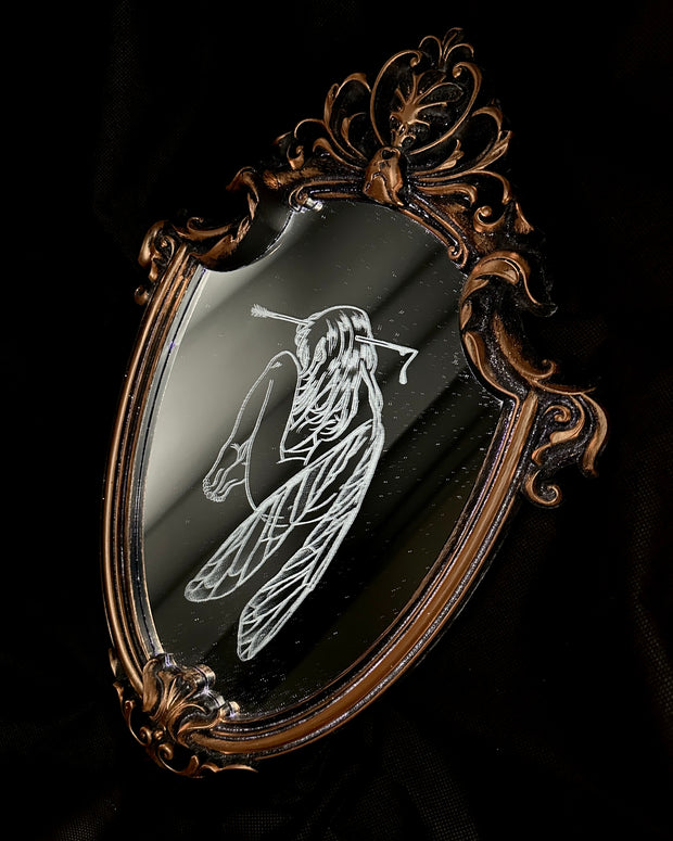 White engraved illustration on a mirror with ornate gold detailing on the outside. Image is of a fairy, with her legs pulled into her chest and an arrow through her head.