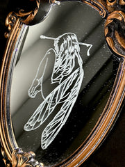 White engraved illustration on a mirror with ornate gold detailing on the outside. Image is of a fairy, with her legs pulled into her chest and an arrow through her head.