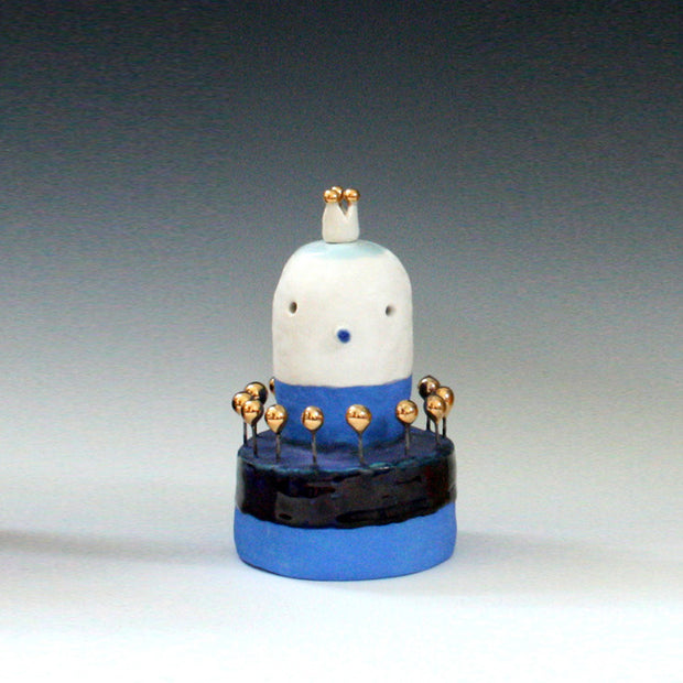 Ceramic sculpture of a white blob character on a blue striped base. Gold balls come out around it on mini stakes.