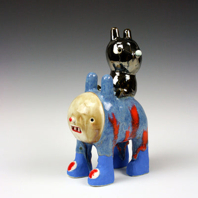 Sculptural piece of a blue quadruped creature with a tan face and shoes. A shiny bunny like creature sits atop its back, as though riding it. 