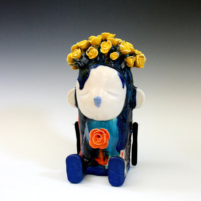 Sculptural piece of a sitting character with dripping blue hair and a yellow flower crown. It has an orange flower growing out of its chest.