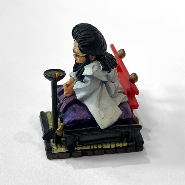 Figure of an old woman with large black hair sitting cross legged on a raised floor seat, atop a tatami mat. Behind her is red guard railing and in front of her is a small black stand.