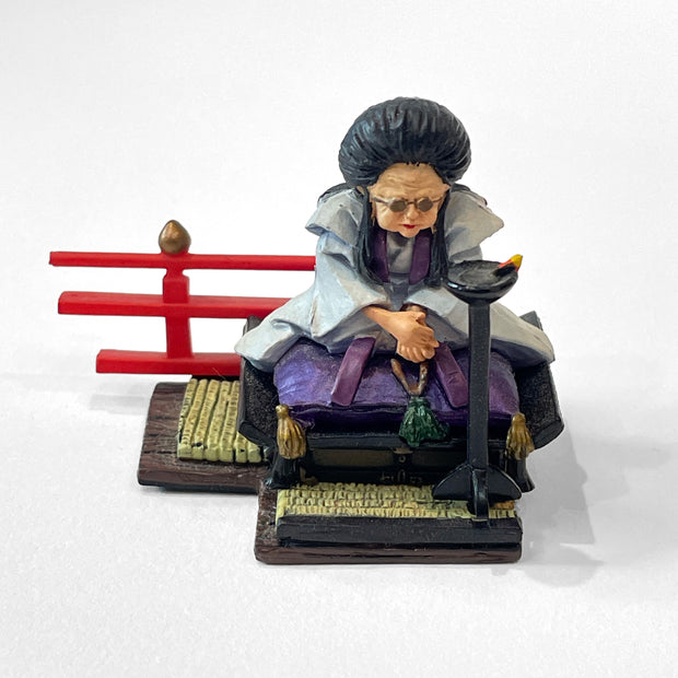 Figure of an old woman with large black hair sitting cross legged on a raised floor seat, atop a tatami mat. Behind her is red guard railing and in front of her is a small black stand.