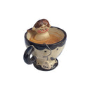 Small ceramic sculpture of a nude girl sitting inside of a tea cup surrounded by coffee colored liquid. A curled wire comes out of the mug with a small tag that reads "tea."