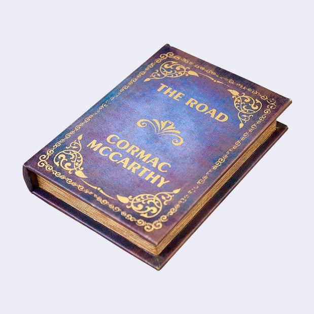 Hardcover book, vintage looking with bluish purple coloring and gold decorative outlining. Title reads "The Road / Cormac McCarthy"