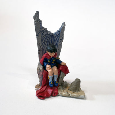 Vinyl figure of a small boy wearing a fancy blue coat with medals attached and matching shorts and shoes. He wears a long red cape and sits atop a stone throne. The throne has a large chunk missing from the top, as though it was shot off.