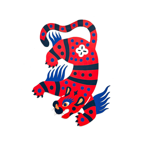 Die cut, brightly painted wooden sculpture of a folklore style tiger, bright red with blue stripes and polka dots. Its body is contorted to mimic movement and it has blue hair on the back of its legs and arms.