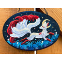 An oval wooden wall hanging featuring cut paper artwork of a white fox with a large flowing red tail on a dark blue night sky background, displayed on a wooden surface.