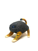 A small sculptural animal figure resembling a cat or tiger, with a dark charcoal textured body, orange-and-black striped legs and tail, and cream paws, photographed on a clean white studio background.