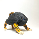 A small sculptural figurine of a four-legged creature with a dark charcoal-black textured body, bright orange and yellow striped legs, and blue eyes, photographed on a plain white studio background.