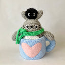 Crochet sculpture of a blue mug with a pink heart. Inside the mug is Totoro with a green scarf. A dust sprite sits atop its head.
