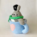 Crochet sculpture of a blue mug with a pink heart. Inside the mug is Totoro with a green scarf. A dust sprite sits atop its head.