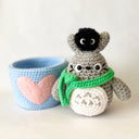 Crochet sculpture of a blue mug with a pink heart. Next to the mug is Totoro with a green scarf. A dust sprite sits atop its head.