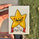 Hand holding an artwork of a star cartoon character with red boots and a mischievous smile, with text that reads "try again!"