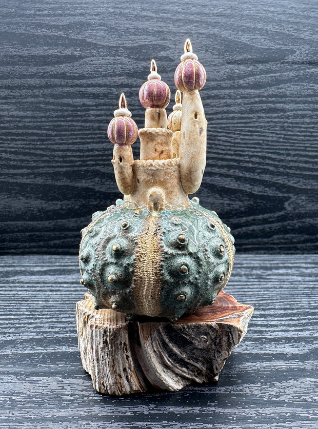 Ceramic sculpture of a rounded piece of green coral of sorts, with a castle sitting atop it with pink rounded tower roofs. Sculpture sits atop of a thick piece of wood.