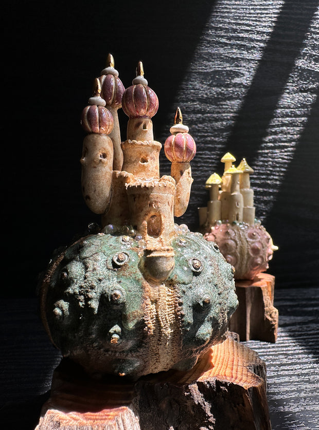 Ceramic sculpture of a rounded piece of green coral of sorts, with a castle sitting atop it with pink rounded tower roofs. Sculpture sits atop of a thick piece of wood.