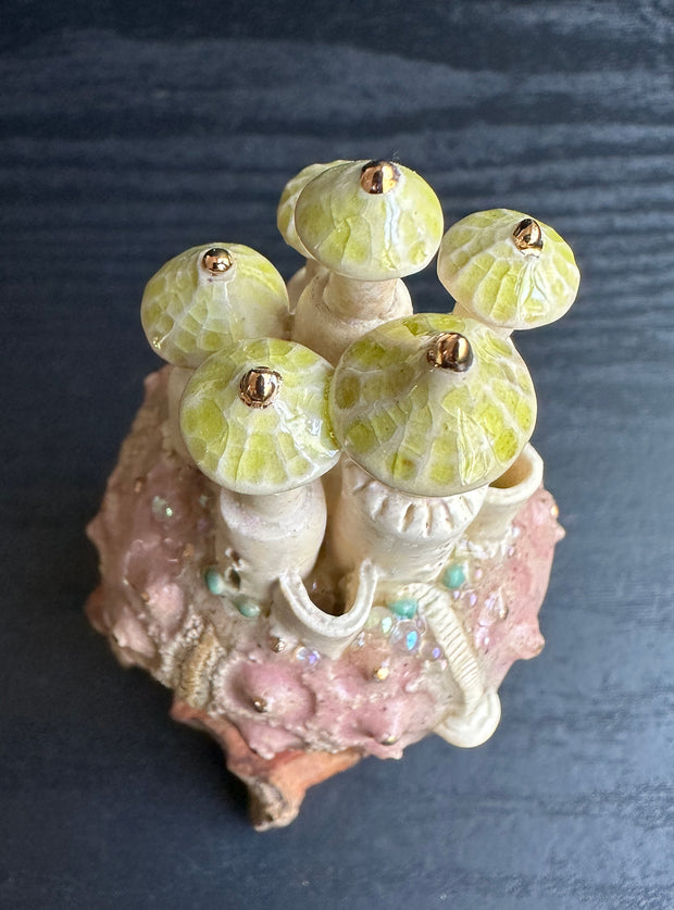 Ceramic sculpture of a rounded piece of pink coral of sorts, with a castle sitting atop it with green peaked tower roofs. Sculpture sits atop of a thick piece of wood.