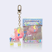 Semi transparent vinyl figure of a unicorn with glitter injection, primarily pink with blue and pink accents. Stars and moons decorate the figure, which is attached to a gold keychain. It stands next to its product packaging.
