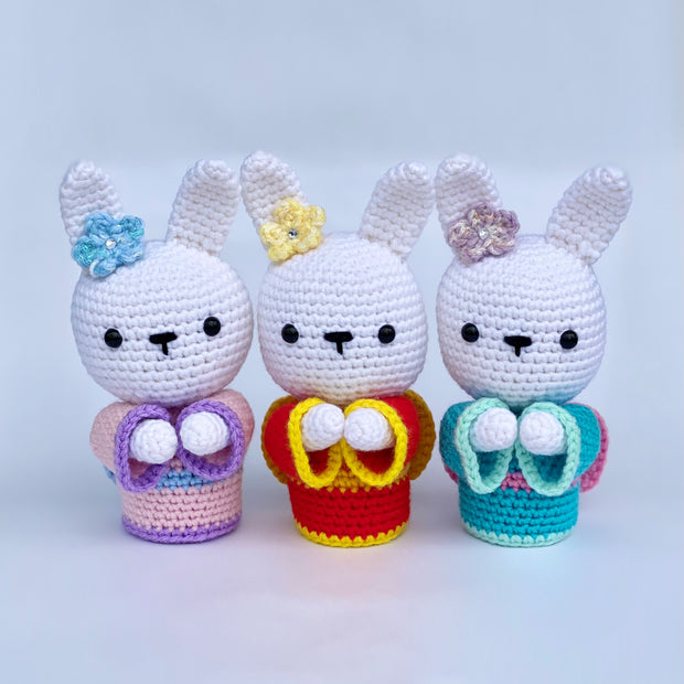 3 crocheted white bunnies, standing up wearing different colored kimonos with matching flowers on their ear. They have simplistic, yet cute faces. Kimono colors are: pink, red, and turquoise.