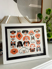 Illustration of 17 dog faces, drawn in orange, black and white. They are done in a very simple, graphic design way. They are of all different breeds and among them is a single dog butt. Framed on a dresser.