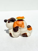 Caterpillar with the face of a Kewpie baby and coloring of a calico cat. Atop its back, is a green plate with an orange fish on it.