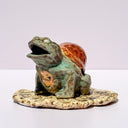 A ceramic turtle figurine with a green glazed body, a reddish-brown shell, and a small painted face on its front, standing on a textured beige base against a plain light background.