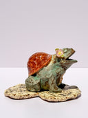 A ceramic turtle figurine with a green glazed body, a reddish-brown shell, and a small painted face on its front, standing on a textured beige base against a plain light background.