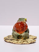 A ceramic turtle figurine with a green glazed body, a reddish-brown shell, and a small painted face on its front, standing on a textured beige base against a plain light background.