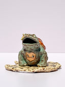 A ceramic turtle figurine with a green glazed body, a reddish-brown shell, and a small painted face on its front, standing on a textured beige base against a plain light background.
