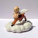 A ceramic figurine of a winged baby figure with a centaur lower half, standing on a speckled white cloud-shaped base against a plain light gray studio background.