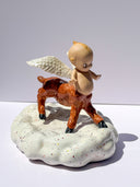 A ceramic figurine of a winged baby figure with a centaur lower half, standing on a speckled white cloud-shaped base against a plain light gray studio background.