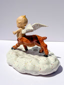 A ceramic figurine of a winged baby figure with a centaur lower half, standing on a speckled white cloud-shaped base against a plain light gray studio background.