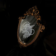 White engraved illustration on a mirror with gold ornate detailing on the exterior. Image is of a wolf with a sheep head atop it with a rope circling around its mouth and behind its head.