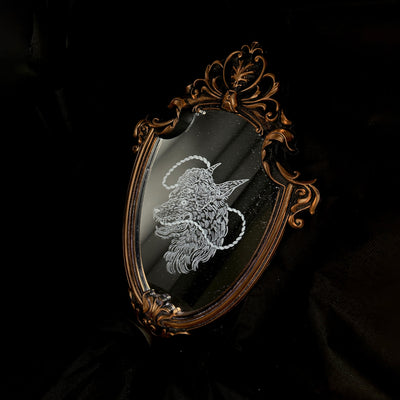 White engraved illustration on a mirror with gold ornate detailing on the exterior. Image is of a wolf with a sheep head atop it with a rope circling around its mouth and behind its head.