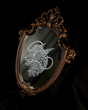 White engraved illustration on a mirror with gold ornate detailing on the exterior. Image is of a wolf with a sheep head atop it with a rope circling around its mouth and behind its head.