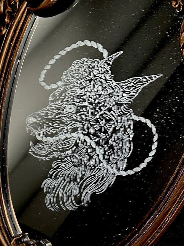 White engraved illustration on a mirror with gold ornate detailing on the exterior. Image is of a wolf with a sheep head atop it with a rope circling around its mouth and behind its head.