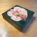 Painting of a brown wolf like dog with a wide snout, curled into a pink flower with semi transparent petals. Background is dark green leaves.