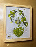 Softly rendered graphite illustration of a woman with very long hair, sitting on the ground in the nude with her eyes closed. Around her are monstera leaves, in the place of her heart and around her lower body. Butterflies fly off the scene. Piece is framed.
