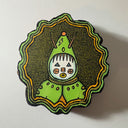 A hand-painted wooden decorative plaque featuring a stylized green creature with a white face, A cross between a clown, a robot and an alien. The figure is set against a textured dark green background with concentric scalloped borders in black, orange, and light green.