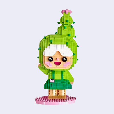 3D sculpture made out of small brick like plastic pieces of a smiling girl, dressed like a cactus and waving. She has a green dress and atop her head is a large blooming cactus.