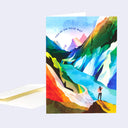 Greeting card with an illustration on the front of a colorful mountain pass with a lake in the middle. A woman stands and looks out at it. Text along the top reads "You're On Your Way"