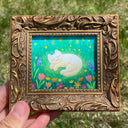 Illustration of a white cartoon cat sleeping in a flower field, drawing is in an ornate bronze frame.