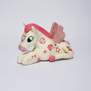 Ceramic figurine of a white horse with a pink mane and pink wings. It sits on the ground with its legs folded under it with red tattoos all over it.