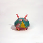 Small ceramic sculpture of a pink bug with large cartoon eyes and a teal blue shell. It has 2 short antenae. 