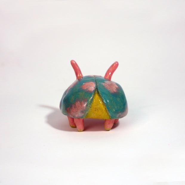 Small ceramic sculpture of a pink bug with large cartoon eyes and a teal blue shell. It has 2 short antenae. 