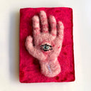 A hand-shaped felt artwork with a green and black eye detail, mounted on a textured bright pink rectangular backing against a plain white background.