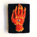 A hand-shaped textile artwork or patch made from bright orange, red, and yellow yarn, mounted on a black rectangular backing with a small blue and white bead at the center, shown against a plain white background.