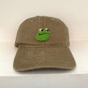 A brown cotton baseball cap featuring a green felt Frog head patch on the front, displayed on a white shelf against a white wall.