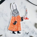 Sticker of a rabbit character in an orange dress with a candle on a marble surface