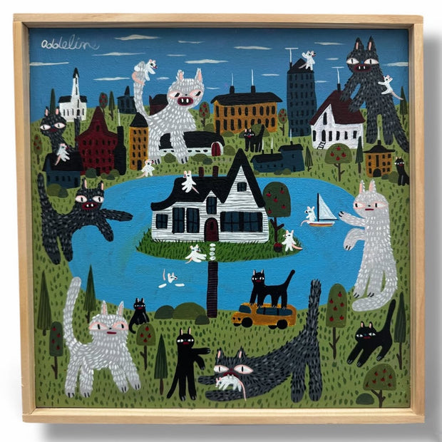 Framed artwork of several stylized cats, very large in size and tormenting a town around a lake. One house sits in the middle of the lake.